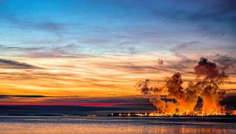 Industrial smoke plumes rising over a Southeast Asian coastline at sunset, highlighting air pollution issues impacting regional health and climate.
