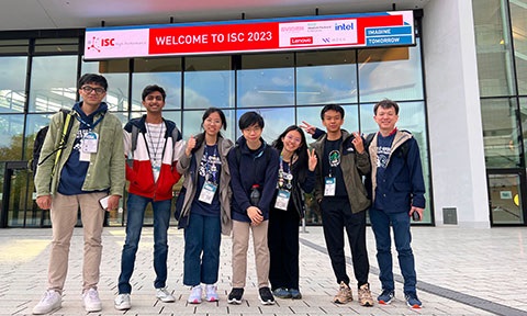 Photo of a group of Uni students and Lecturer posing in front of a glass building with welcome banner.