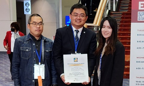 Photo of a professor holding an award certificate standing between 2 postgraduates.