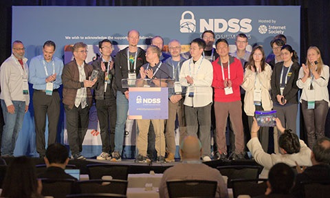 A thumbnail photo of a group of researchers at NDSS conference stage.