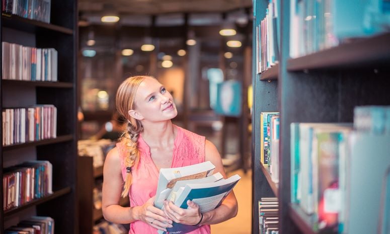 Woman holding books looking at bookshelves in library