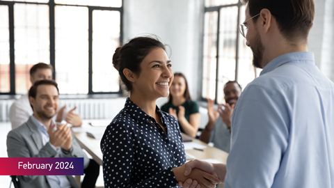 Employee getting promotion shaking hands with boss and receiving praises from colleagues in the background with February 2024 text on bottom left