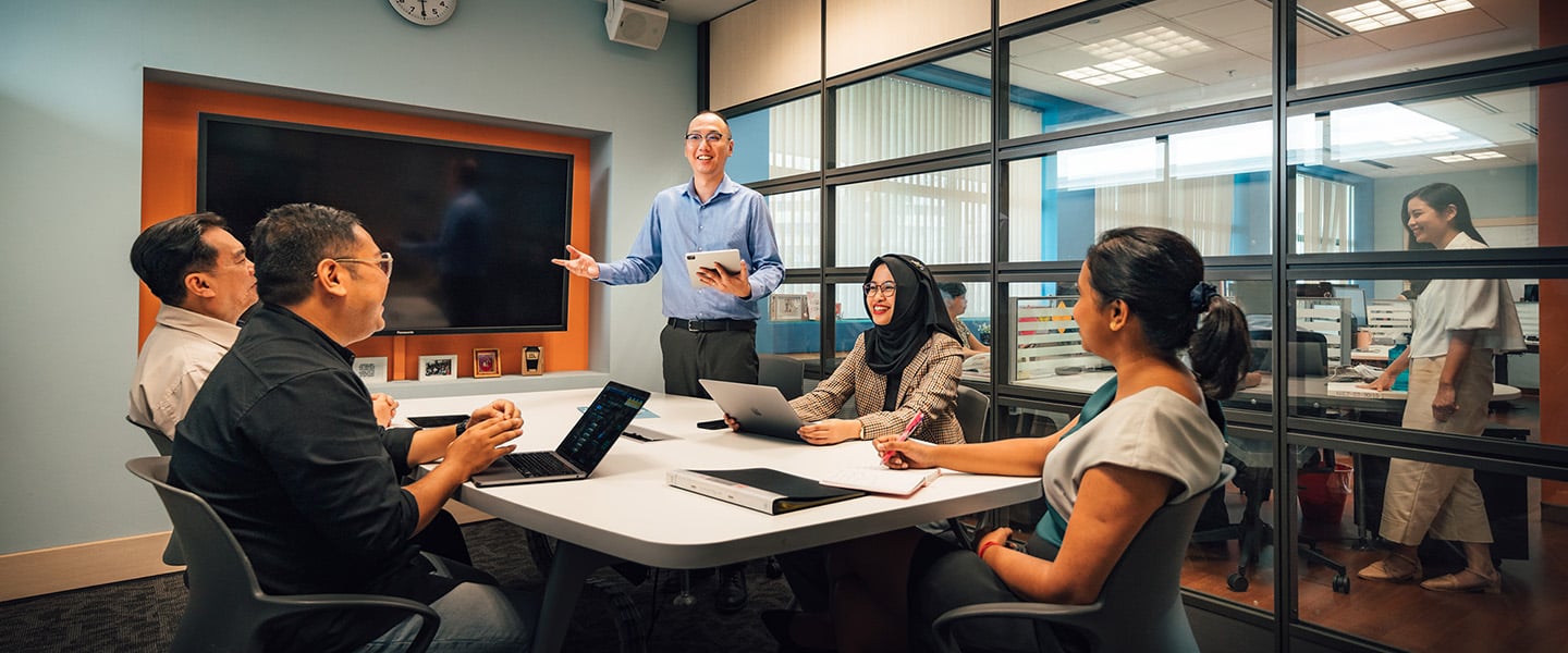 Business Conference Room Man In Blue Shirt Holding Ipad Presenting To A Group Of Colleagues Having A Discussion