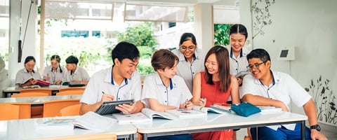 Female Teacher in Orange Dress with Group of 5 Secondary Students in Uniform Studying with Textbooks