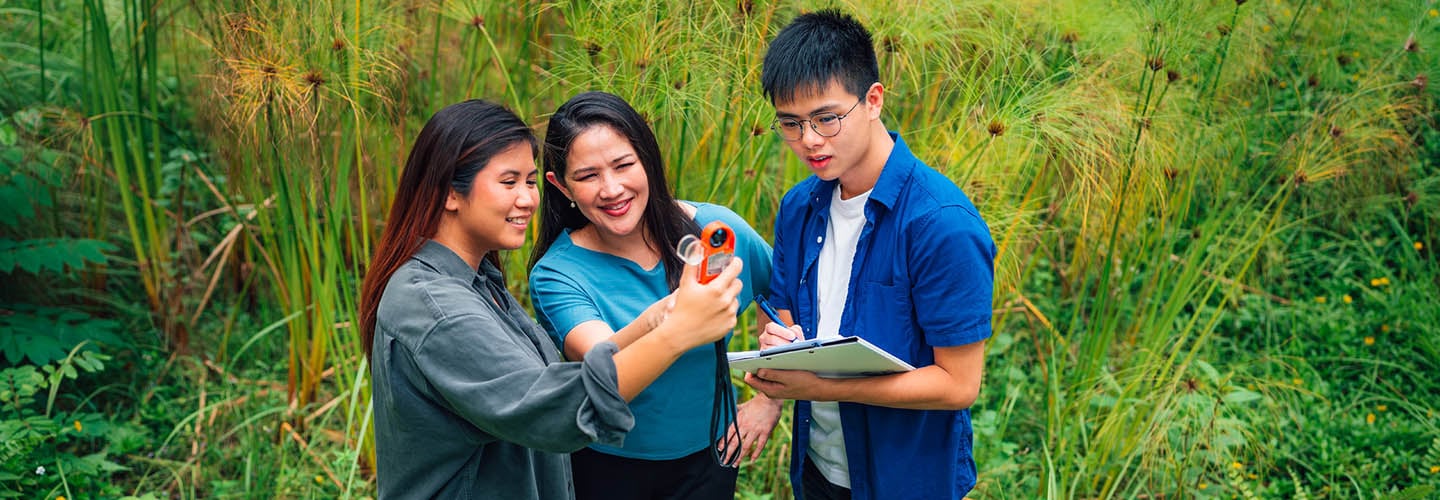 Outdoor Open Field Science Trip Female Teacher In Blue Top Holding An Orange Device. 2 Young Adult Students. Holding A Clipboard Doing An Experiment