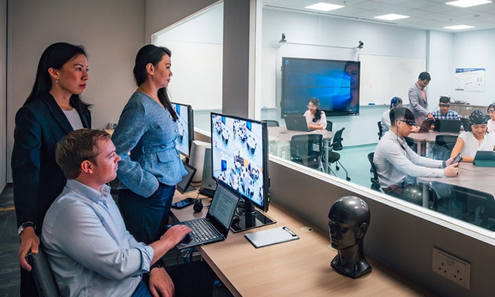 EEG Brain Caps SoLEC Classroom with Caucasian Man Looking At Computer Screen Monitoring Students Inside Of Classroom Behind The Glass Window (720x432px)
