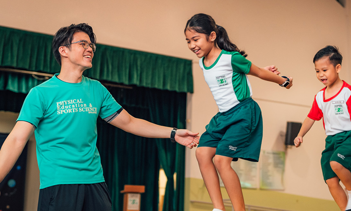 Male PE Teacher Instructing Group Of 4 Primary School Kids Jumping On Gymnastics Equipment Wideshot 2 (720x432px)