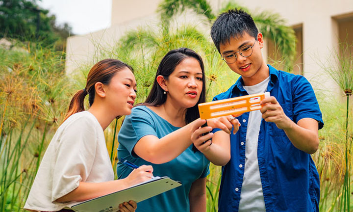 Outdoor Open Field Science Trip Female Teacher In Blue Top Holding A Yellow Scale. 2 Young Adult Students. Holding A Clipboard Doing An Experiment (720x432px)