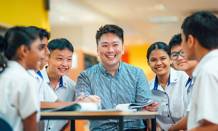 Smiling Male Teacher in Library with Group of 7 Secondary Students in Uniform (720x432px)