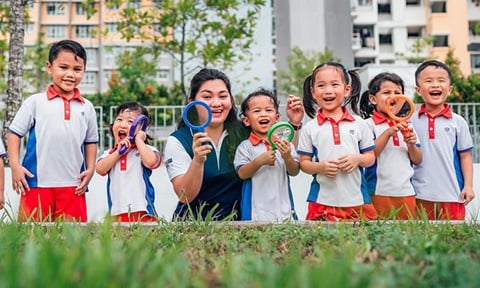 Toy Magnifying Glass Kindergarten Students And Teacher Outside On A Grassy Field Looking Straight Ahead All Smiles Wideshot (720x432px)