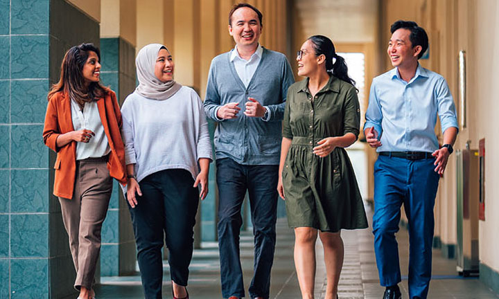 Walking Shot Tall Male Teacher In Grey Cardigan Along NIE Campus With 4 Other Young Adult Students In Casual Businesswear (720x432px)