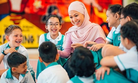 Yong Malay Lady Teacher With Pink Tudung With Group Of 8 Secondary School Students Smiling and Laughing Infront of Art Wall Wideshot (720x432px)