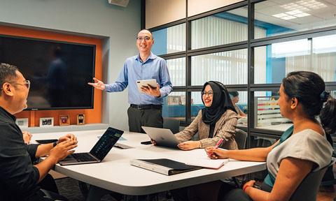 iPad Bald Man In Blue Shirt Holding Ipad Business Conference Room Presenting To A Group Of Colleagues Having A Discussion (480x288px)