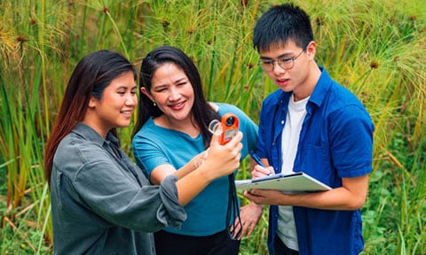 Outdoor Open Field Science Trip Female Teacher In Blue Top Holding An Orange Device. 2 Young Adult Students. Holding A Clipboard Doing An Experiment (480x288px)