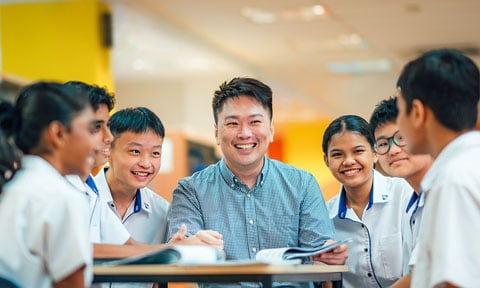 Smiling Male Teacher in Library with Group of 7 Secondary Students in Uniform (480x288px)
